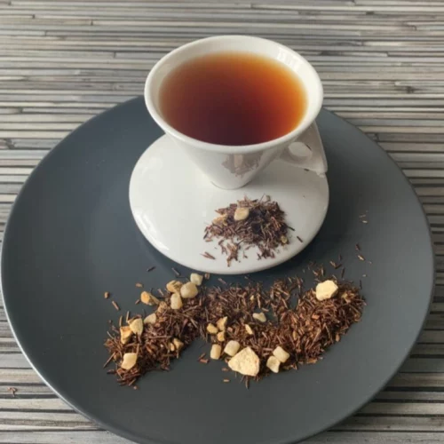 Rotbusch Ingwer Zitrone mit Rotbuschtee, Ingwerstücken und Zitronenschalen, kupferfarbene Tassenfarbe in weißer Teetasse auf gestreiftem Hintergrund