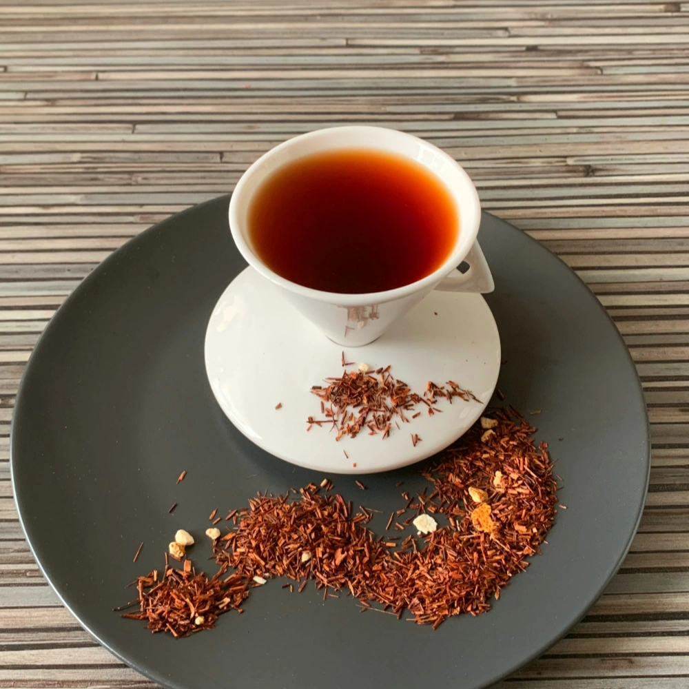 Rotbusch Leuchtende Sonne mit Rooibos und Orangenschalen, kupferfarbene Tassenfarbe in weißer Teetasse auf gestreiftem Hintergrund