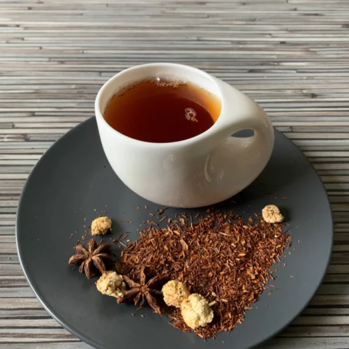 Rotbusch Orientalische Nacht Vanille Zimt mit Rotbuschtee, Sternanis und Zimtstücke, kupferfarbene Tassenfarbe in weißer Teetasse auf gestreiftem Hintergrund.