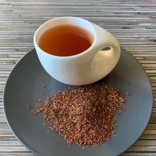 Rotbusch pur in weißer Tasse aufgegossen, lose Rooibos-Blätter auf grauem Teller, gestreifter Hintergrund