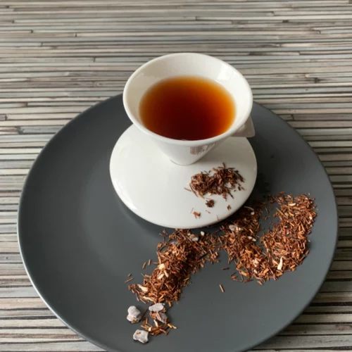 Rotbusch Schoko Sahne Trüffel in weißer Tasse, lose Teemischung mit Schokoladenplättchen und Kokosraspeln im Teller auf gestreiftem Untergrund