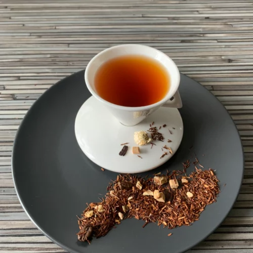 Rotbusch Tiramisu Mascarpone in weißer Tasse, lose Teemischung mit Schokoladenplättchen und Krokantstücken im Teller auf gestreiftem Untergrund