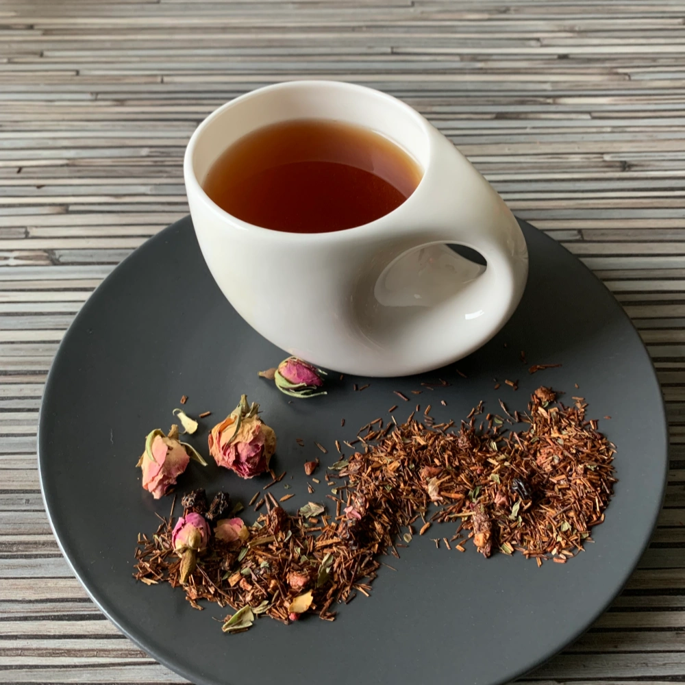 Rotbusch Traubenernte in weißer Tasse, lose Teemischung mit Rotbusch und pinken Rosenknospen im Teller auf gestreiftem Untergrund