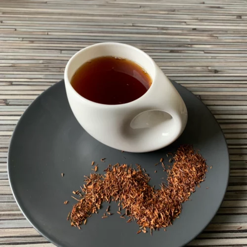 Rotbusch Windhuk Vanille in weißer Tasse, lose Teemischung im Teller auf gestreiftem Untergrund