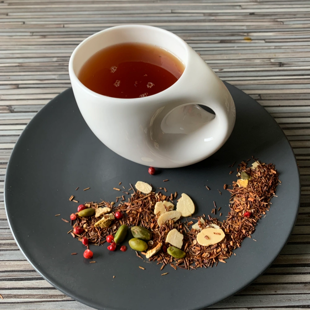 Rotbusch Wintertee in weißer Tasse, lose Teemischung mit Orangenschalen, Mandeln und rosa Pfefferkörnern im Teller auf gestreiftem Untergrund