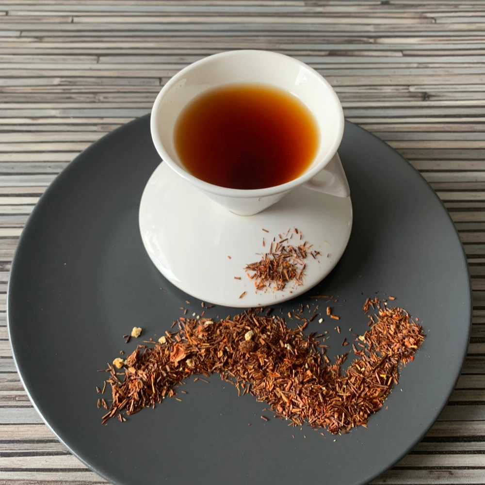 Rotbusch Zauber Afrikas Granatapfel Grapefruit kupferfarben in weißer Tasse, lose Teemischung mit Granatapfelkernen im Teller auf gestreiftem Untergrund