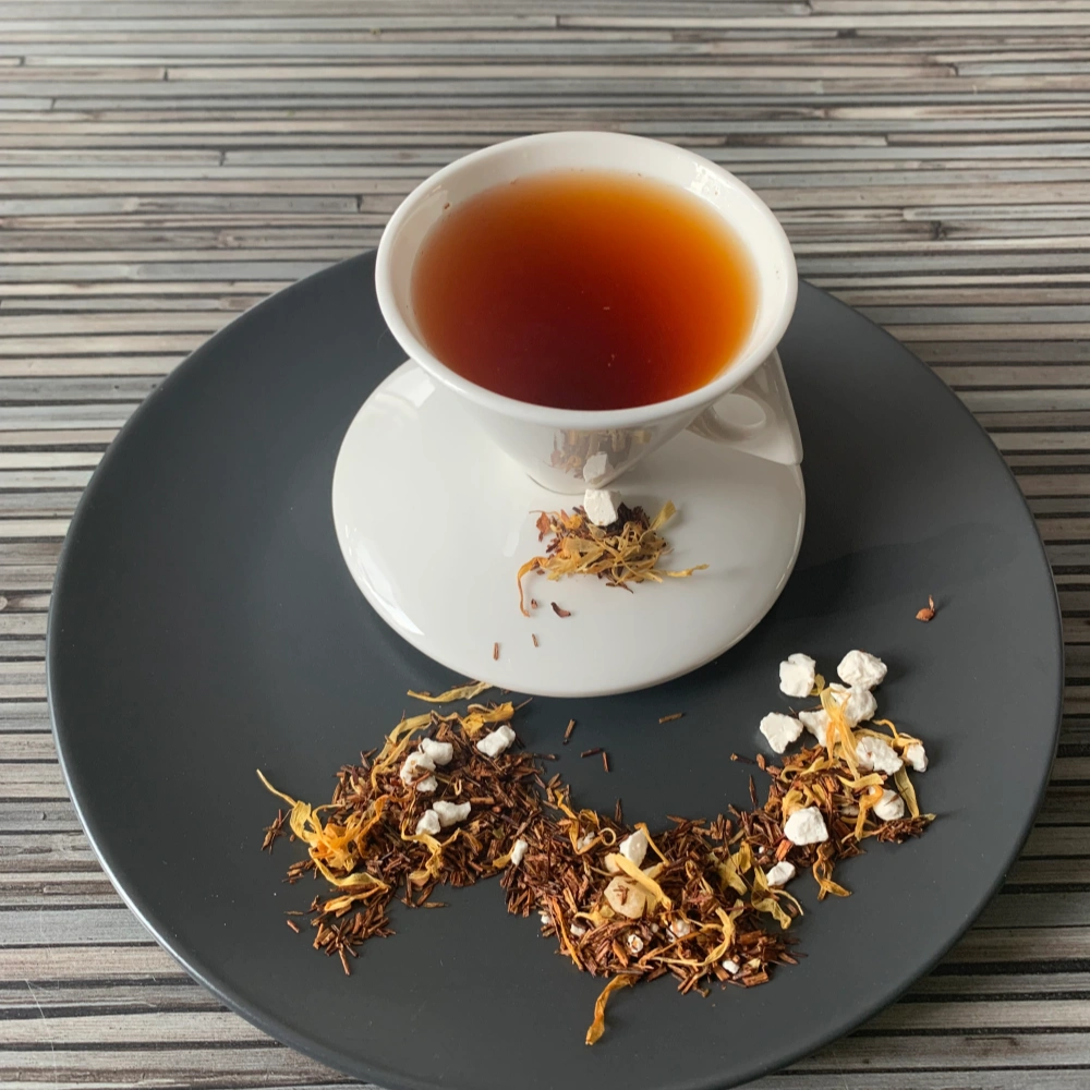 Rotbusch Ananas Zitronen Sorbet mit Rotbuschtee, Ananaswürfeln, Zitrusfruchtstücken und Ringelblumenblüten und heller, kupferfarbener Tassenfarbe in weißer Teetasse auf gestreiftem Hintergrund