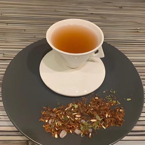 Rotbuschtee Episkey mit warm brauner Farbe, Schokoladenplättchen und Minzblättern in weißer Tasse, auf gestreiftem Holzhintergrund