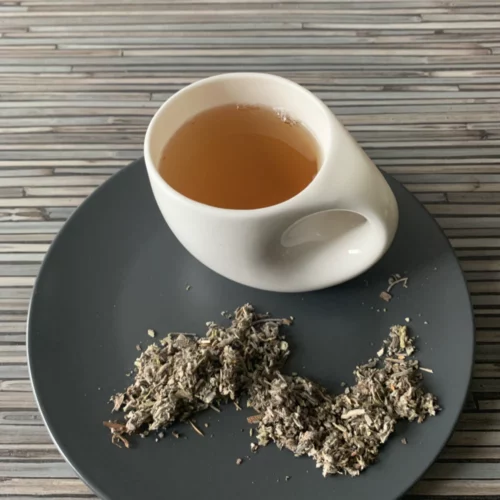 Salbeiblätter geschnitten Tee in weißer Tasse mit warmem Aufguss, auf grauem Teller mit losem Tee, arrangiert auf gestreiftem Holzhintergrund