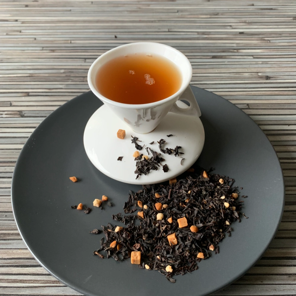 Schwarztee Salziges Karamell in weißer Tasse auf dunklem Teller auf gestreifter Holzplatte, loser Tee mit Karamell- und Mandelstücken daneben