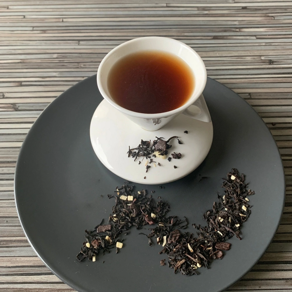 Schwarztee Schokkoko in weißer Tasse auf dunklem Teller auf gestreifter Holzplatte, loser Tee mit Schokoladen- und Kokosstücken daneben
