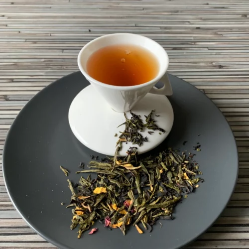 Schwarz-Grüntee Des Kaisers 7 Kostbarkeiten in weißer Tasse auf dunklem Teller auf gestreifter Holzplatte, loser Tee mit Blütenblättern daneben