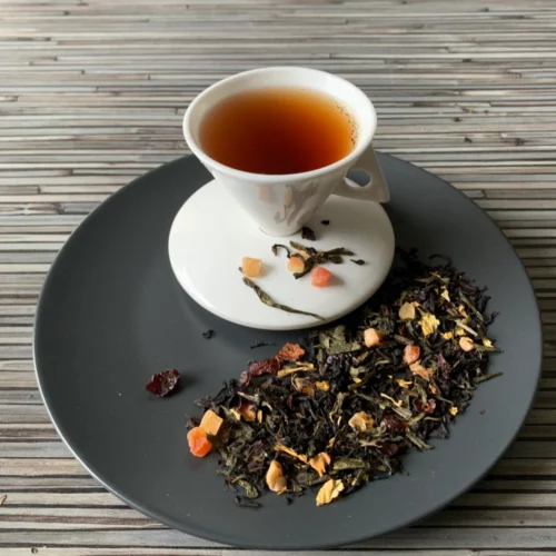 Schwarz-Grüntee Zaubermond Erdbeer in weißer Tasse auf dunklem Teller auf gestreifter Holzplatte, loser Tee mit Blüten und Fruchtstücken daneben