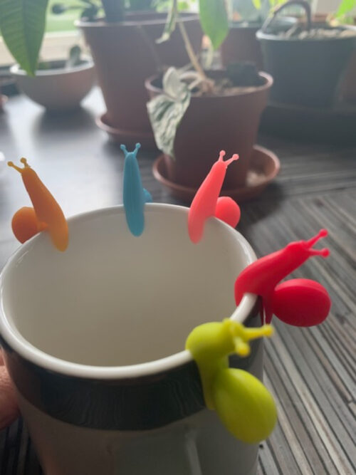 Teebeutelhalter Schnecke aus Silikon, hängend an einer grauen Tasse auf gestreiftem Hintergrund.