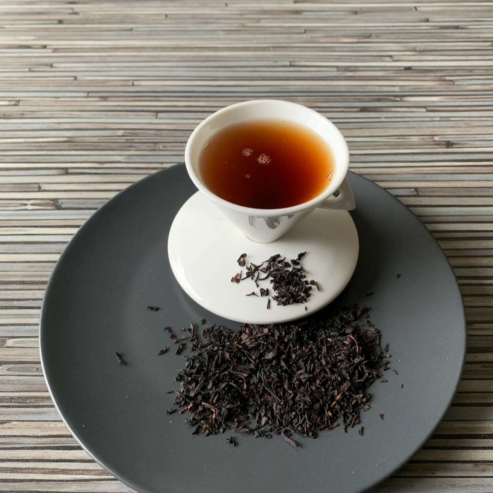 Schwarztee Vanille mit Vanillestücken in weißer Tasse auf dunklem Teller auf gestreifter Holzplatte, loser Tee mit Vanillestücken daneben