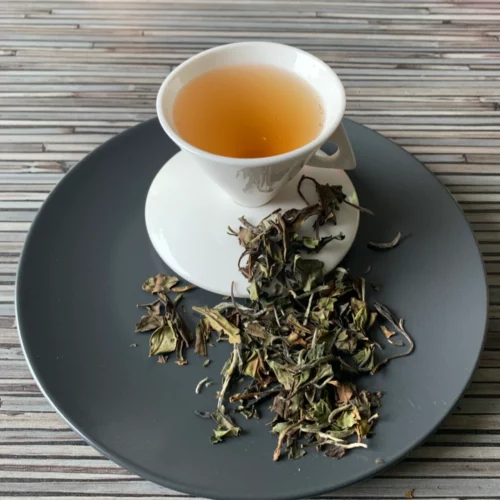 Weißer Tee China Pai Mu Tan in weißer Tasse auf dunklem Teller auf gestreifter Holzplatte, loser weißer Tee daneben