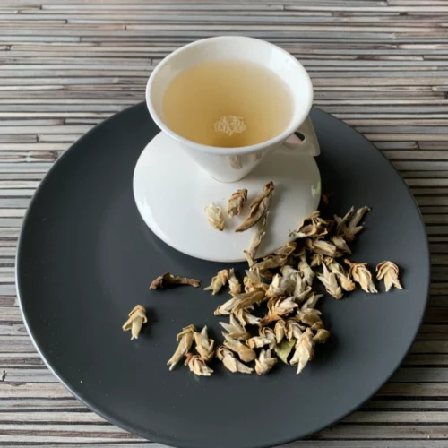 Weißer Tee China Yunnan Silver Bud Ya Bao in weißer Tasse auf dunklem Teller auf gestreifter Holzplatte, lose helle Teeknospen daneben
