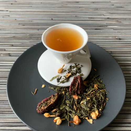 Grün-Weißer Tee Elegance Vanille Jasmin in weißer Tasse auf dunklem Teller auf gestreifter Holzplatte, loser Tee mit Jasminblüten und Erdbeerstücken daneben