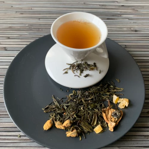 Grün-Weißer Tee Sonnige Passion in weißer Tasse auf dunklem Teller auf gestreifter Holzplatte, loser Tee mit Mangowürfeln, Ananasflocken und Rosenknospen daneben